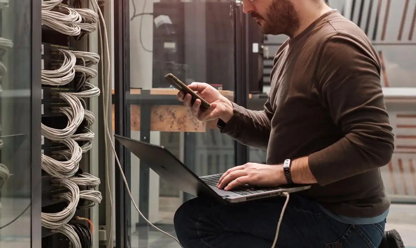 man looking at phone with computer while working on server